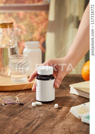 On the wooden table, trays containing water bottles, water glasses and medicine boxes are decorated. hand of woman is preparing to pick up a bottle of medicine unlabeled. Scene for product advertising On the wooden table, trays containing water bottles, water glasses and medicine boxes are decorated. hand of woman is preparing to pick up a bottle of medicine unlabeled. Scene for product advertising 117316772