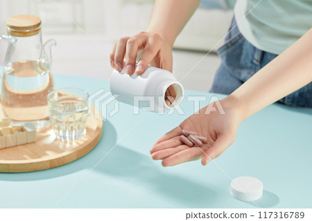 Hands of woman are pouring out pills in preparation for taking them. Trays containing water bottles, water glasses and medicine boxes are decorated on blue table 117316789