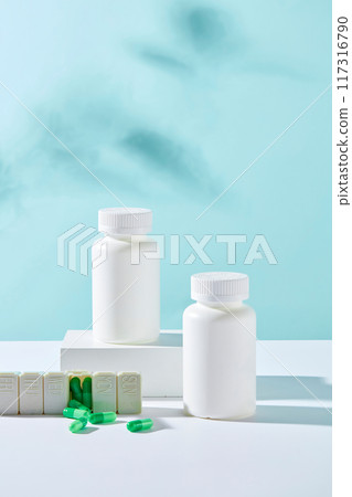 Front view of two white bottles unlabeled displayed on white podiums with medicine daily box and green capsules on blue background. Minimal style for advertising medicine product 117316790