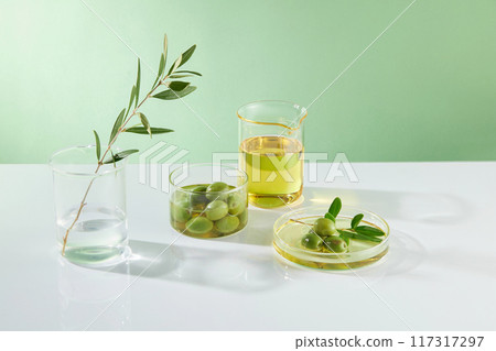 Beaker containing oil, fresh olives and branches decorates on a green background. Minimal scene for advertising product with ingredient from olive extract Beaker containing oil, fresh olives and branches decorates on a green background. Minimal scene for advertising product with ingredient from olive extract 117317297