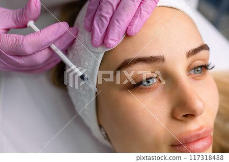 Close up of hands of young cosmetologist injecting botox in female face. She is standing and smiling. The woman is closed her eyes with relaxation 117318488