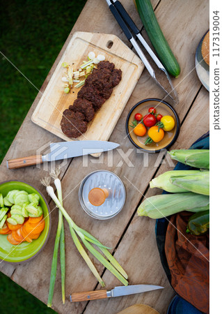Top view of cutting board with sharp knife, chopped vegetables and grilled burger patties. Preparing food for an outdoor barbecue. grilling setup. 117319004