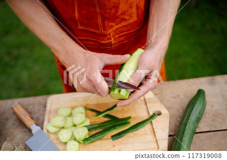 Close up of man holding sharp knife, peeling cucumber. Preparing vegetables for an outdoor barbecue. Close up of man holding sharp knife, peeling cucumber. Preparing vegetables for an outdoor barbecue. 117319008