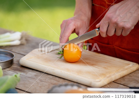 Close up of man holding sharp knife, slicing juicy red tomato. Preparing vegetables for an outdoor barbecue. Close up of man holding sharp knife, slicing juicy red tomato. Preparing vegetables for an outdoor barbecue. 117319018