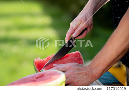 Close up of man holding sharp knife, slicing juicy red watermelon. Preparing food for an outdoor barbecue. Close up of man holding sharp knife, slicing juicy red watermelon. Preparing food for an outdoor barbecue. 117319029