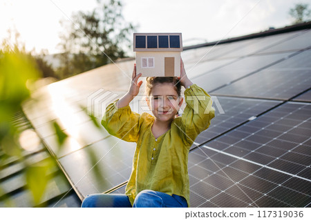Cute girl holding model of house with solar panels, sitting on rooftop solar or photovoltaic system. Sustainable future for next generation. 117319036