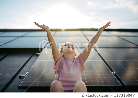 Cute girl on roof with solar panels, standing with open arms, hands up in the air. Rooftop solar or photovoltaic system. Sustainable future for next generation. 117319047