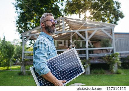 Man looking at solar panel in arms, standing in garden. Sustainable lifestyle and green household. Man looking at solar panel in arms, standing in garden. Sustainable lifestyle and green household. 117319115