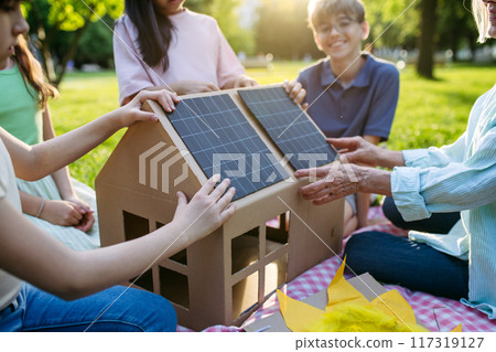 Children learning about renewable energy and solar panels during sustainable education class outdoors, using cardboard model of house wit solar panel on roof. 117319127