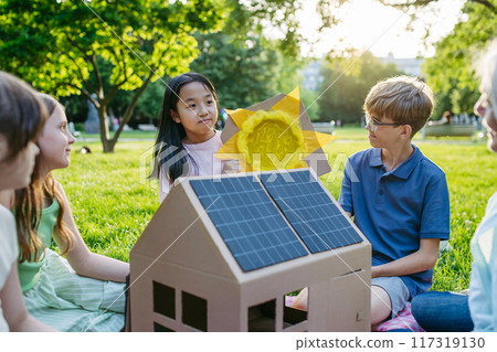 Children learning about renewable energy and solar panels during sustainable education class outdoors, using cardboard model of house wit solar panel on roof. Children learning about renewable energy and solar panels during sustainable education class outdoors, using cardboard model of house wit solar panel on roof. 117319130