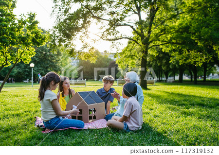 Children learning about renewable energy and solar panels during sustainable education class outdoors, using cardboard model of house wit solar panel on roof. Children learning about renewable energy and solar panels during sustainable education class outdoors, using cardboard model of house wit solar panel on roof. 117319132