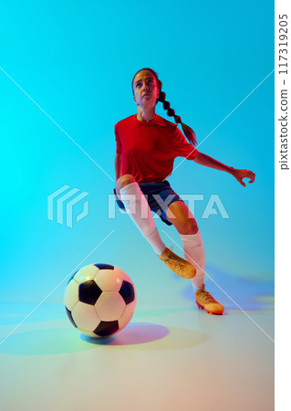 Low angle view of young female soccer player focused on ball training dribbling technique in neon light against colorful gradient background. 117319205