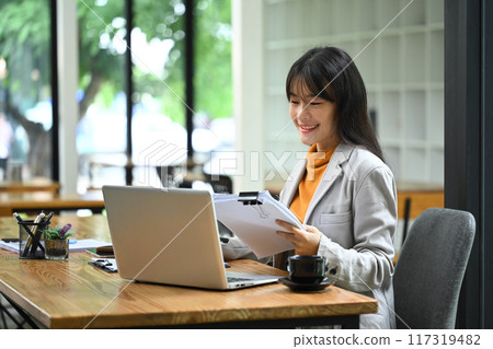 Young businesswoman in stylish suit checking financial document and using laptop at office desk. 117319482