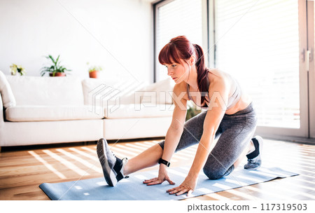 Beautiful redhead woman's home fitness routine. Home workout on gym mat in living room, sporty woman wearing sportswear. 117319503