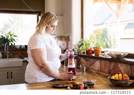 Overweight woman in the kitchen, making fresh smoothie in blender. Choosing healthy food, making smoothie at home. Overweight woman in the kitchen, making fresh smoothie in blender. Choosing healthy food, making smoothie at home. 117319515