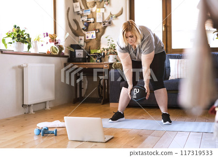 Online fitness training for overweight woman exercising at home, using weighted medicine ball. Workout in living room. Online fitness training for overweight woman exercising at home, using weighted medicine ball. Workout in living room. 117319533
