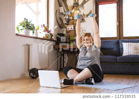 Online fitness training for overweight woman exercising at home with dumbbells, looking at laptop. Workout in living room. Online fitness training for overweight woman exercising at home with dumbbells, looking at laptop. Workout in living room. 117319535