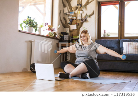 Online fitness training for overweight woman exercising at home with dumbbells, looking at laptop. Workout in living room. 117319536