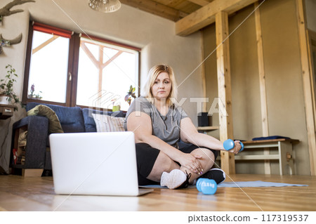 Online fitness training for overweight woman exercising at home, looking at laptop. Workout in living room. 117319537