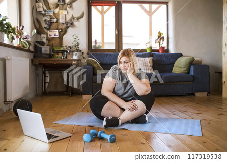 Online fitness training for overweight woman exercising at home, looking at laptop. Workout in living room. Online fitness training for overweight woman exercising at home, looking at laptop. Workout in living room. 117319538