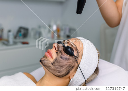 Close-up of cosmetologist's hand making hardware carbon peeling by young pretty customer woman in safety glasses headdress lying on couch in beauty salon. Facial cleaning concept. Cosmetology Services Close-up of cosmetologist's hand making hardware carbon peeling by young pretty customer woman in safety glasses headdress lying on couch in beauty salon. Facial cleaning concept. Cosmetology Services 117319747