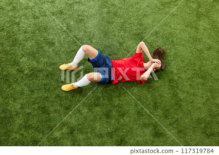 Girl, soccer player lies on football field, twisted in pain. Player violation against opponent. Moment of foul. Aerial view Girl, soccer player lies on football field, twisted in pain. Player violation against opponent. Moment of foul. Aerial view 117319784