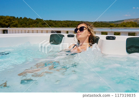 Woman wearing sunglasses and swimsuit relaxing in outdoor hot tub with bubbling water and enjoying sunny day. Female resting in outdoor jacuzzi with beautiful natural landscape 117319835