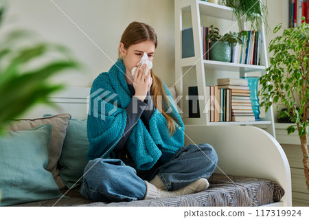Young sick woman sitting under warm blanket sneezing coughing in handkerchief 117319924