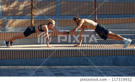 Fit man and woman training in sunny morning, doing plank hand tap, combining strength, balance, and teamwork in their outdoor workout routine. 117319932