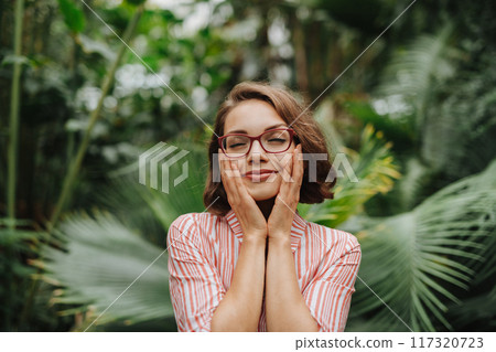 Beautiful woman standing in botanical garden, in the middle of lush green foliage, holding her face. 117320723