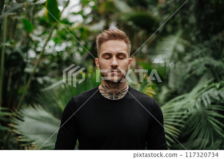 Handsome man with tattoo standing in botanical garden, in the middle of lush green foliage, looking at camera. Handsome man with tattoo standing in botanical garden, in the middle of lush green foliage, looking at camera. 117320734