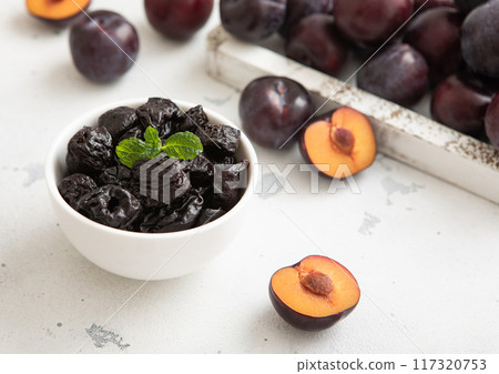 Sweet prunes in glass bowl with ripe plums in wooden box on light kitchen background.Macro. 117320753