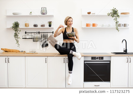 Fit woman eating healthy salad in modern white kitchen Fit woman eating healthy salad in modern white kitchen 117323275