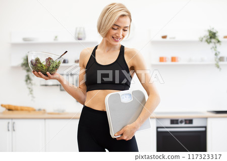 Healthy woman holding salad bowl and weighing scale in kitchen 117323317
