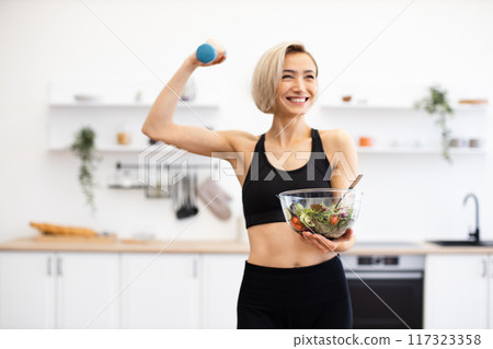 Fit woman holding salad and weight in modern kitchen Fit woman holding salad and weight in modern kitchen 117323358