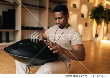 Relaxed African man playing on modern musical instrument Hand pan, Vadjraghanta, metal tongue drum sitting on floor at home. Silence calm filling room by sounds of music from musical instrument. 117323669