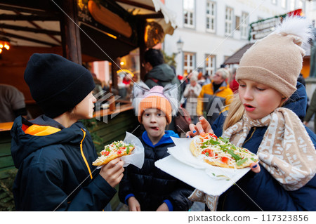 Three cute children kids wear warm winter clothing enjoy freshly made street food langos bread at german Christmas outdoor market  winter festival. Festive atmosphere holiday spirit concept 117323856