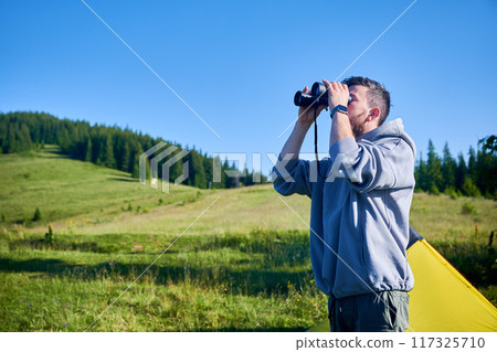 Man stands in grassy field, looking through binoculars, wearing gray hoodie. On background gentle hill covered in lush greenery and line of tall pine trees under clear blue sky. 117325710