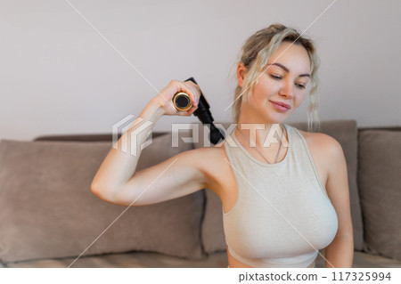 Girl at home holding a therapeutic percussion vibrating massage gun in her hand and massaging her back muscles Sports recovery concept after a workout.  117325994