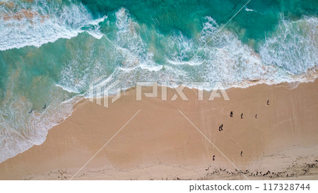 Aerial view of turquoise waves gently crashing onto a sandy beach. The oceans clear water, mixed with white foam, creates a tranquil 117328744