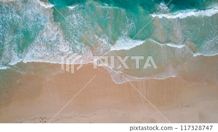 Aerial view of turquoise waves gently crashing onto a sandy beach. The oceans clear water, mixed with white foam, creates a tranquil 117328747