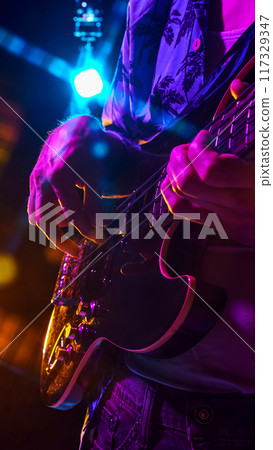Fingers expertly pluck guitar strings under neon lights, capturing essence of live performance with burst of color and sound. 117329347