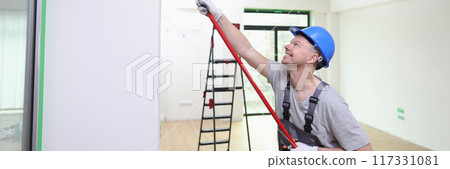 Smiling decorator in helmet painting wall with roller brush 117331081