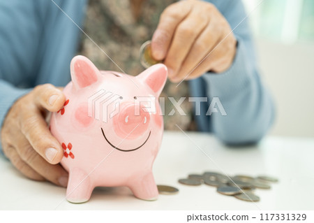 Asian elderly woman putting coin into pink piggy bank for saving money and insurance, poverty, financial problem in retirement. 117331329