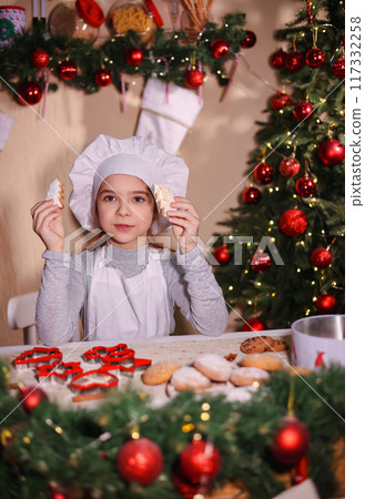 A cheerful girl in a chef's hat shows Christmas gingerbread cookies in the shape of a Christmas tree. 117332258