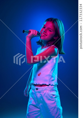 Young positive, emotional girl in white clothes singing in microphone against blue studio background in neon light. Creative hobby 117332334