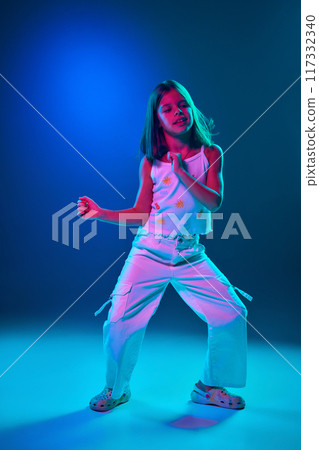 Full-length image of young girl in casual clothes dancing, having fun against blue studio background in neon light. Entertainment 117332340
