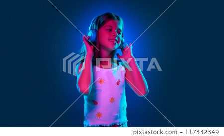 Young 9 years old girl listening t music in headphones, enjoying song against blue studio background in neon light Young 9 years old girl listening t music in headphones, enjoying song against blue studio background in neon light 117332349
