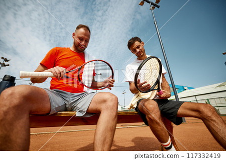 Two men, tennis player sitting on bench on outdoor court and preparing for the game, checking rackets, getting ready for intense training Two men, tennis player sitting on bench on outdoor court and preparing for the game, checking rackets, getting ready for intense training 117332419
