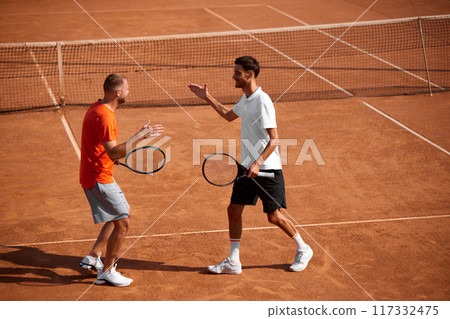 Two emotional happy men, tennis players in motion on clay court showing support and respect to each other, shaking hands after golf game 117332475